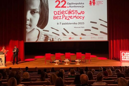 scena z pustymi czerwonymi krzesłami a nad nia ekran na którym wyświetla się slajd z nazwą konferencji Dzieciństwo bez przemocy
