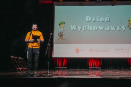 Na scenie mężczyzna w żółtej bluzie coś czyta. W tle ekran z napisem Dzień Wychowawcy oraz rysunkami postaci małego chłopca i starszego pana oraz logotypami Miasta st. Warszawy, Centrum Wspierania Rodzin "Rodzinna Warszawa" i "Terminalu Klutury"