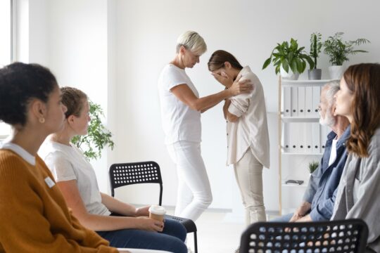 Sala w której siedzi kilka osób i stją dwie kobiety. Jedna ukrywa twarz w rękach druga wydaje sie ją pocieszać.