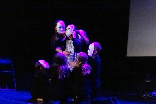 Grupa pięciu postaci w maskach na scenie. Ciemne tło i fioletowe światło tworzą atmosferę tajemniczości.